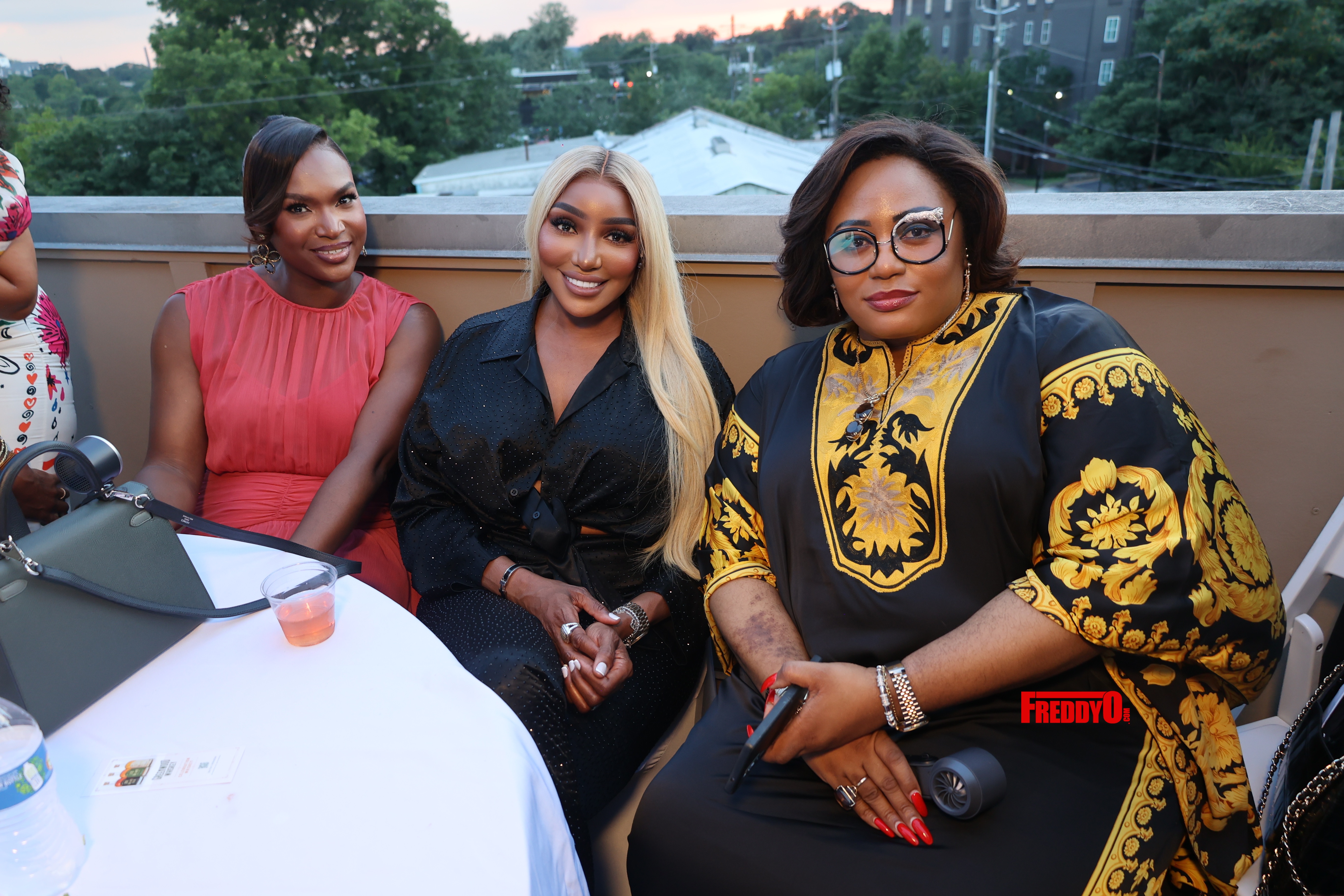 Three women posing together outdoors, one wearing a red dress, one in a black outfit, and one in a yellow and black patterned dress.