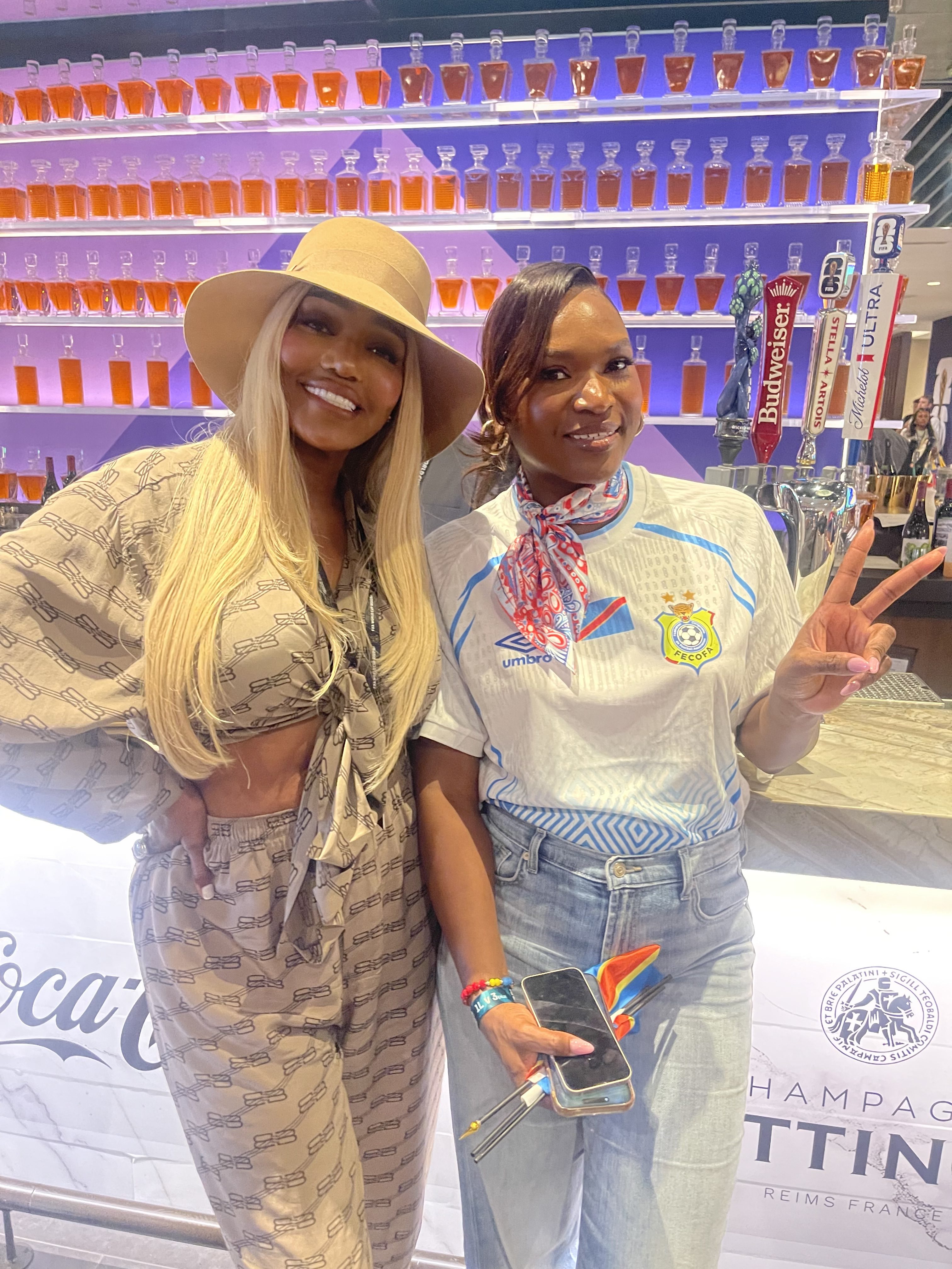 Two women smiling and posing in front of a colorful display of liquor bottles, one wearing a yellow hat and the other wearing a white jersey with the Peco logo.