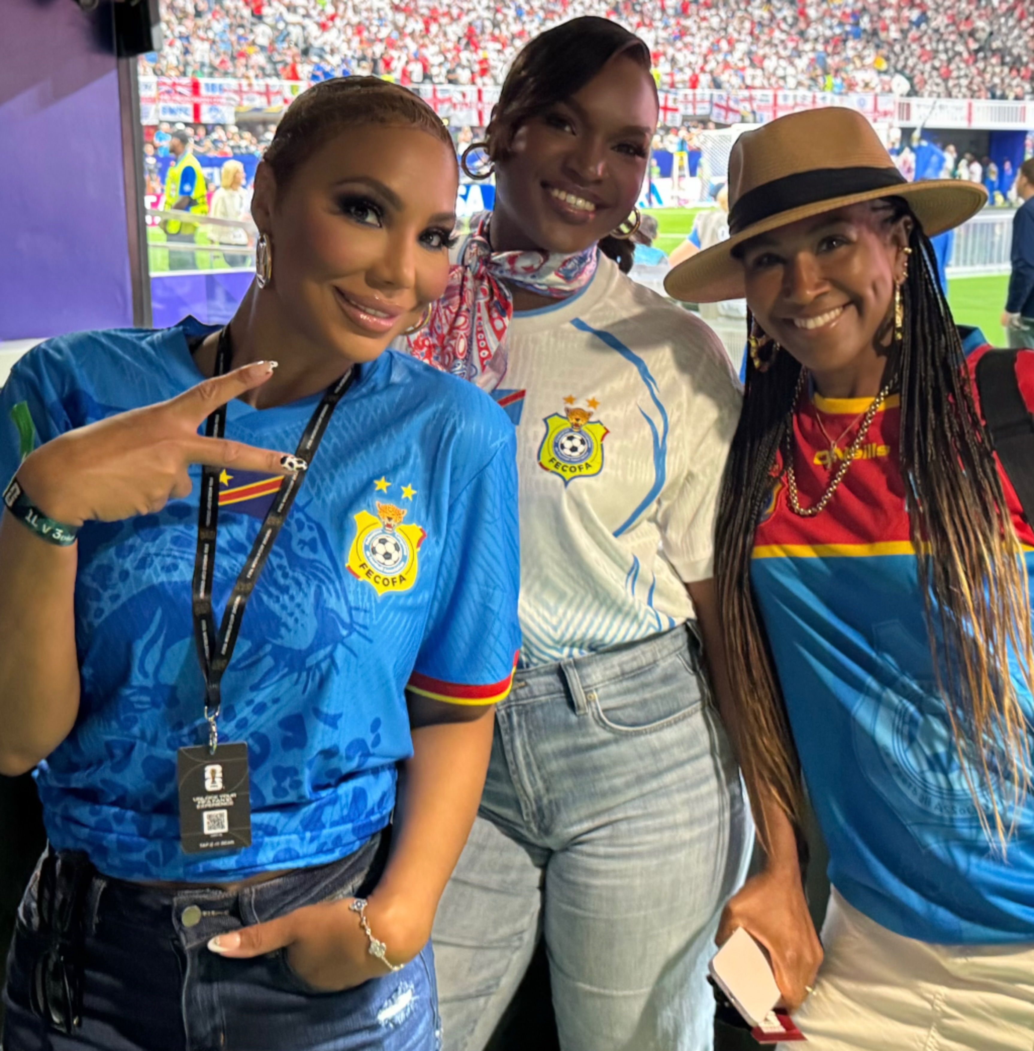 Three smiling women wearing soccer jerseys and hats, posing together in a crowded stadium.