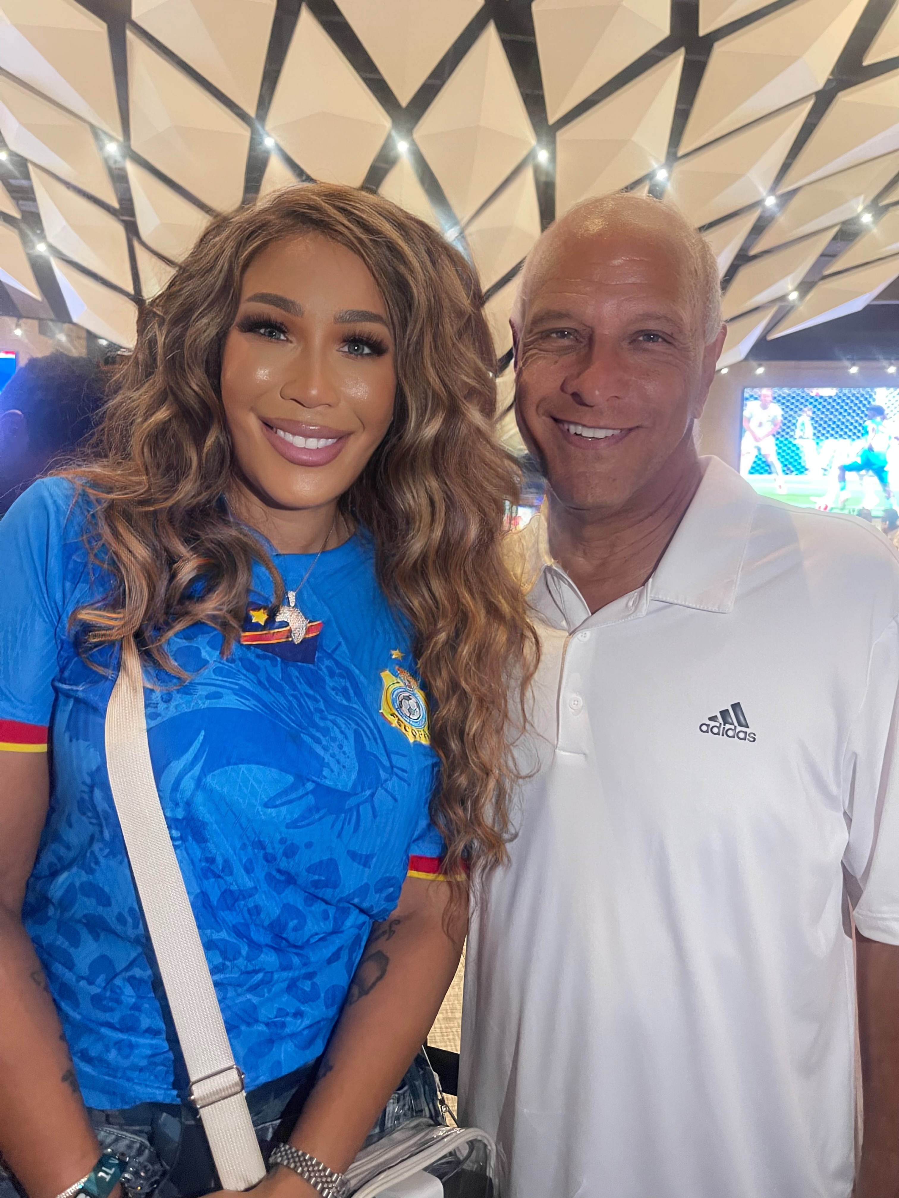 Two people smiling and posing together in front of a patterned background, one wearing a blue sports jersey and the other wearing a white shirt.