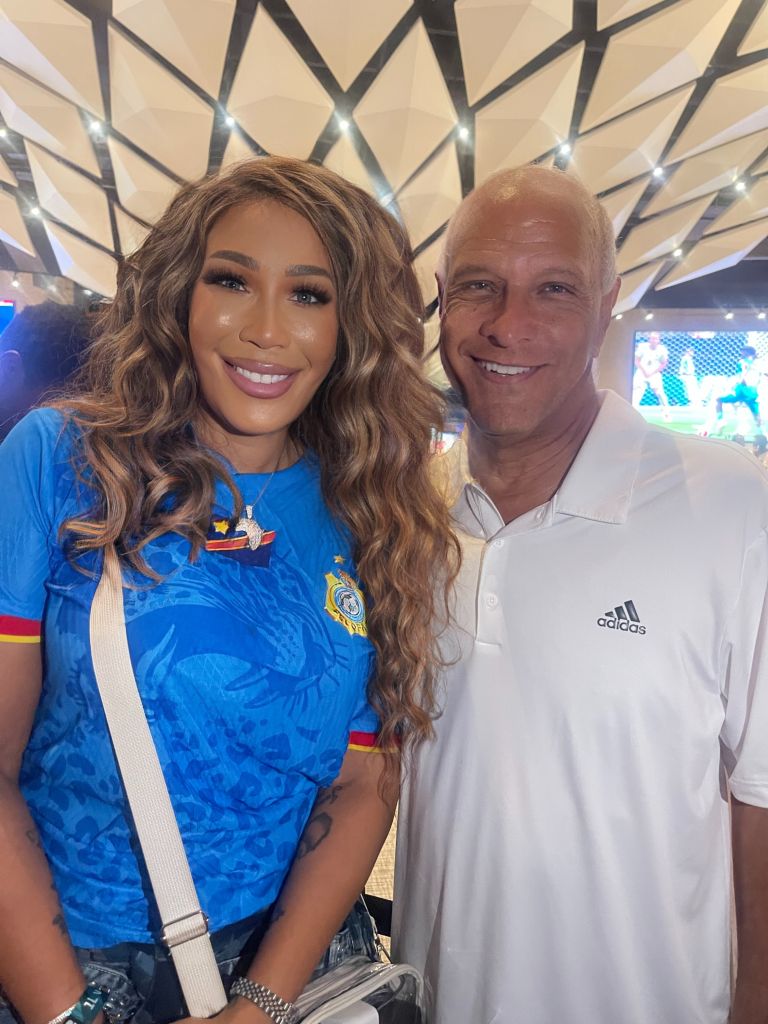 Two people smiling and posing together in front of a patterned background, one wearing a blue sports jersey and the other wearing a white shirt.
