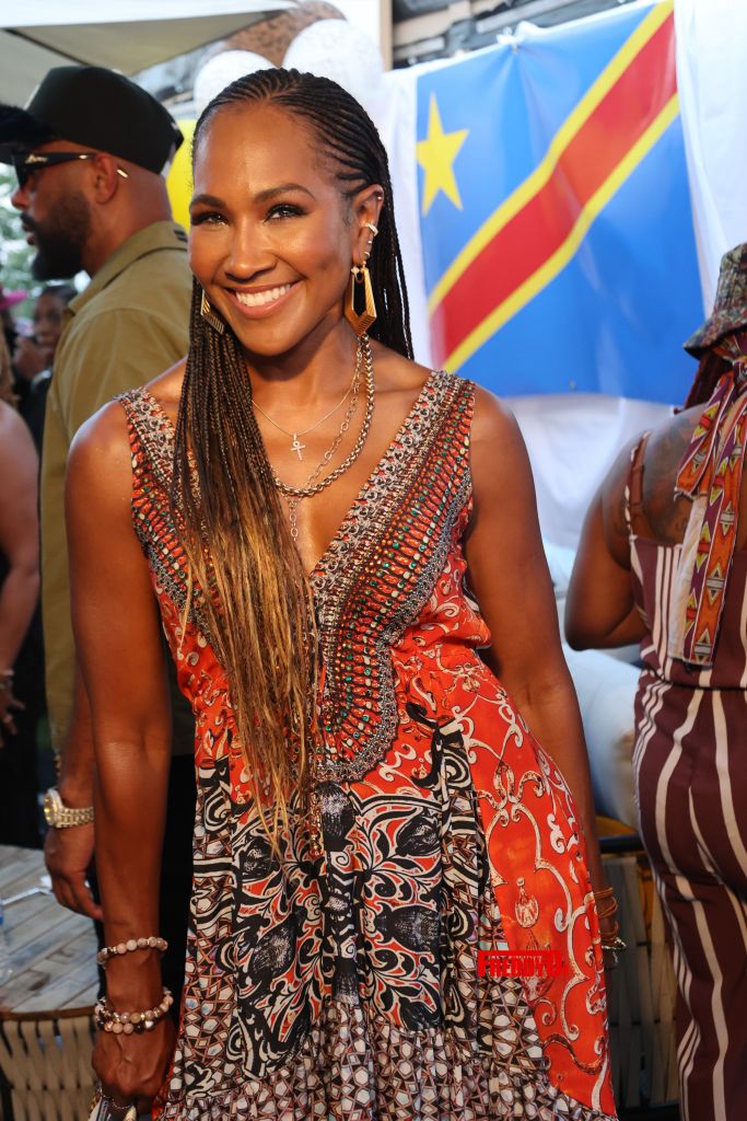 A smiling woman with braided hair wearing a colorful patterned dress and jewelry, standing in front of a Democratic Republic of the Congo flag.