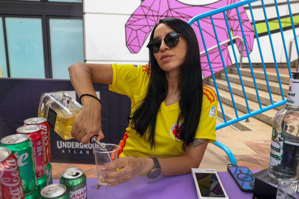A young woman wearing sunglasses and a yellow t-shirt with the Underground Asian logo, sitting at a table with various canned drinks and a mobile phone.