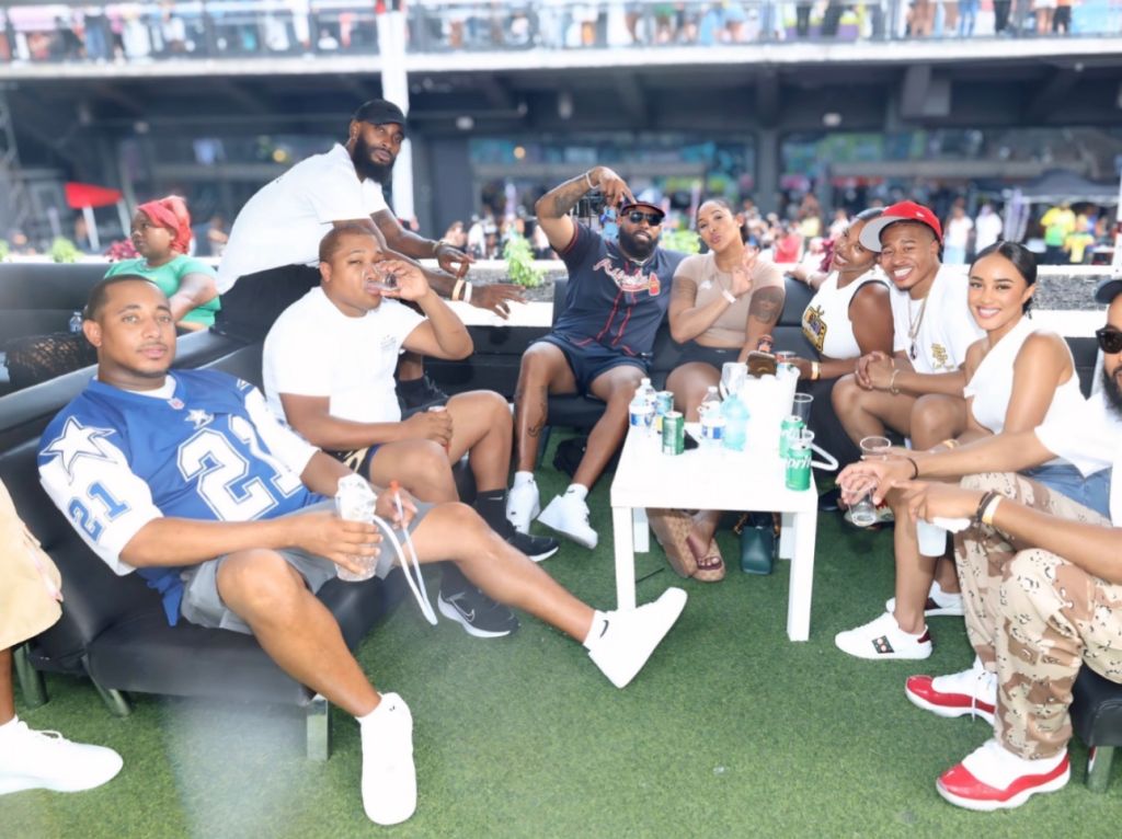 A group of people, including several men and women, sitting together and socializing outdoors on a grassy area with a stadium or arena in the background.