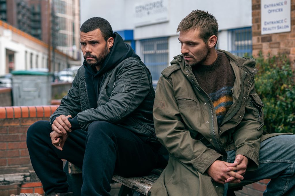 Two men sitting on a bench in an urban setting, wearing jackets and looking pensive.