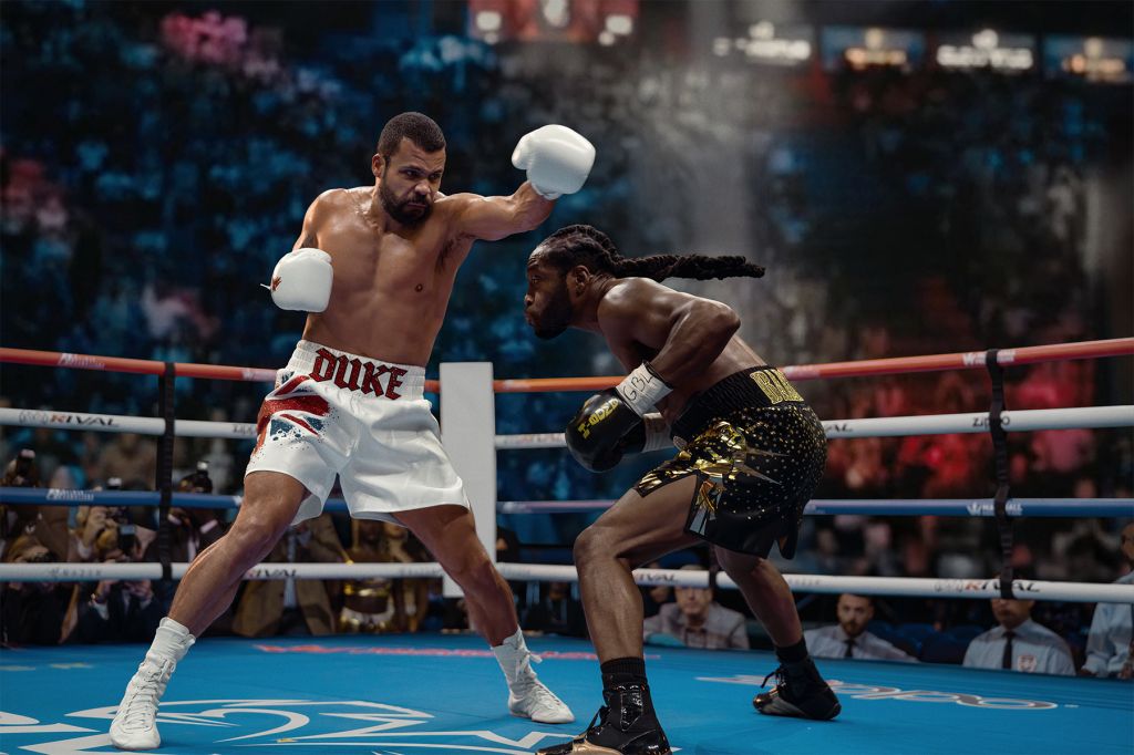 Two boxers in a boxing ring, one wearing white trunks with "DUKE" printed on them, the other wearing gold and black trunks, engaged in a boxing match.