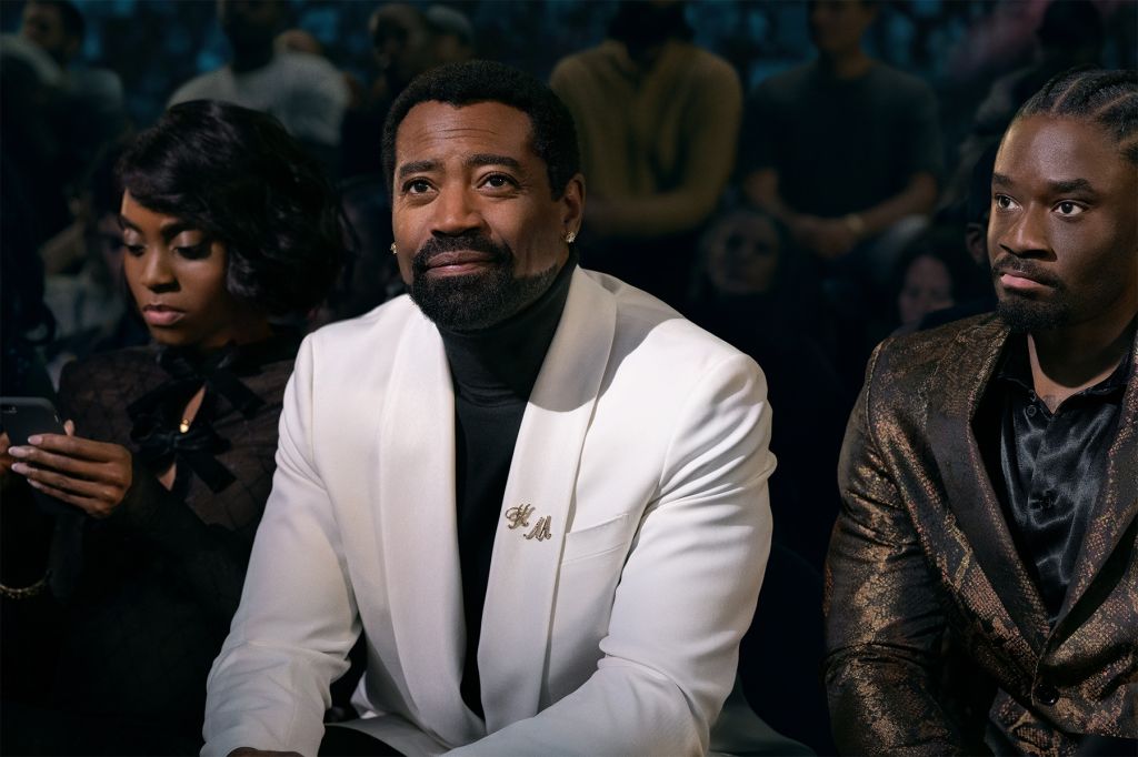 Three Black men sitting in a dimly lit room, one wearing a white suit jacket with "H.M." embroidered on it.