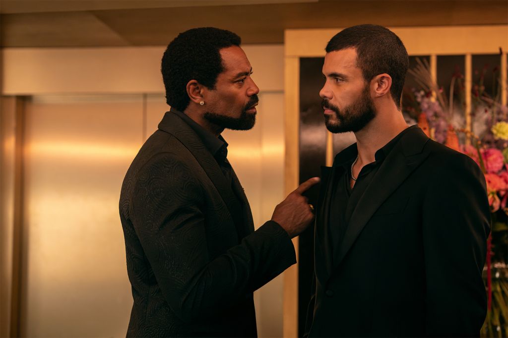 Two men in black suits facing each other in a dimly lit room with floral arrangements in the background.