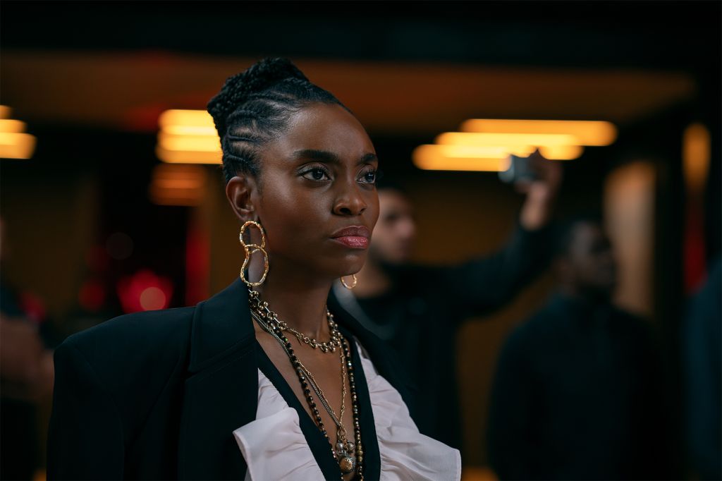 A close-up portrait of a young Black woman with braided hair wearing a black blazer and gold jewelry, looking intently at the camera against a dimly lit background.