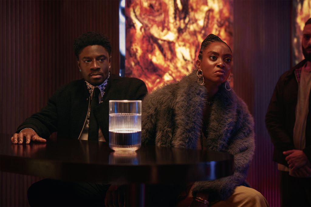 Two people, a man and a woman, sitting at a table in a dimly lit room, with a large, fiery image projected behind them.