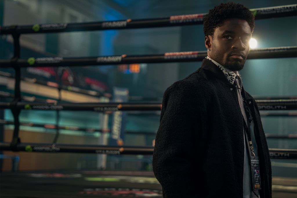 A man with a beard wearing a black coat stands in a dimly lit room with shelves of boxing equipment behind him.