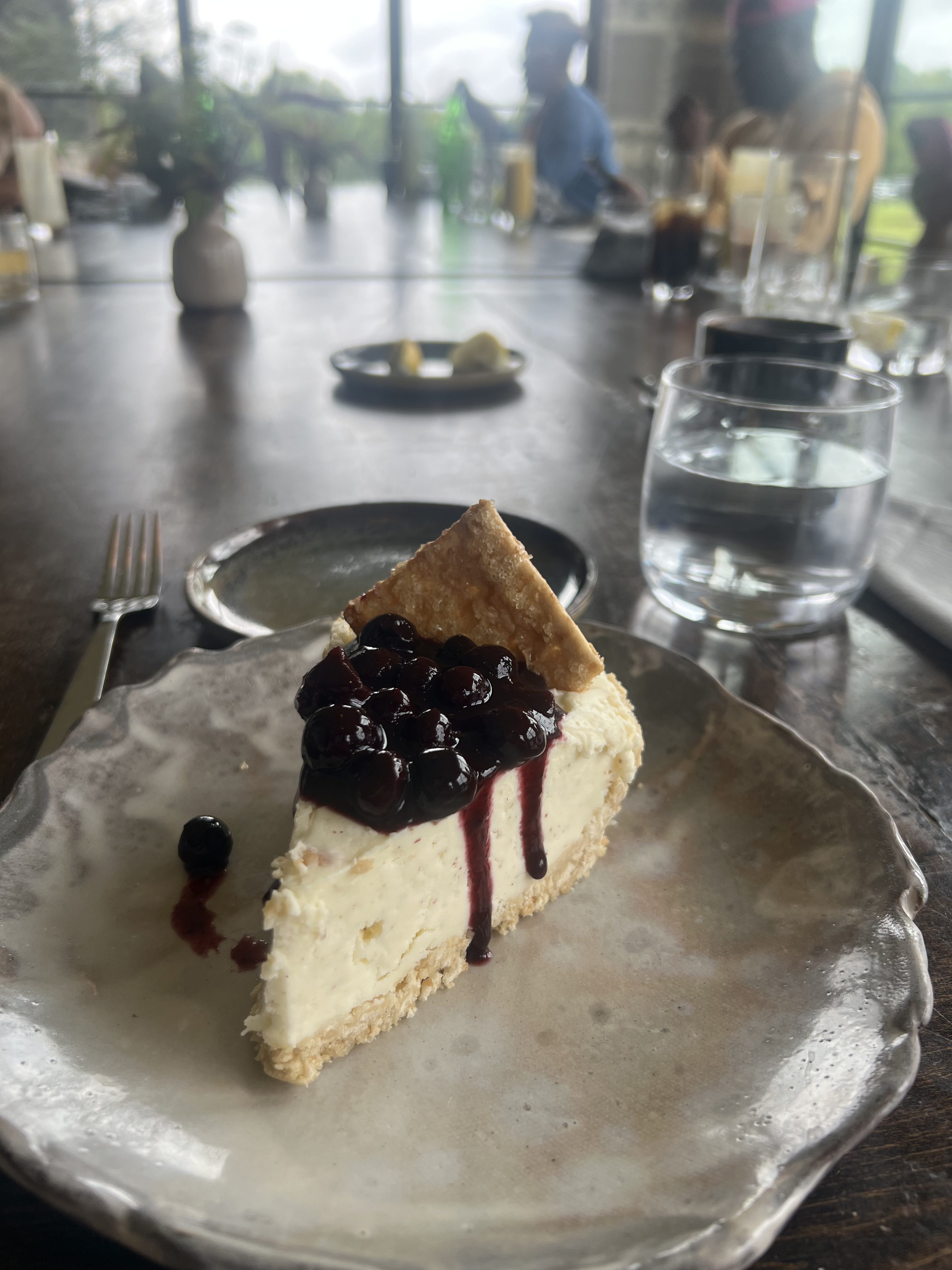 A slice of cheesecake topped with blueberries on a plate, with a glass of water in the background.
