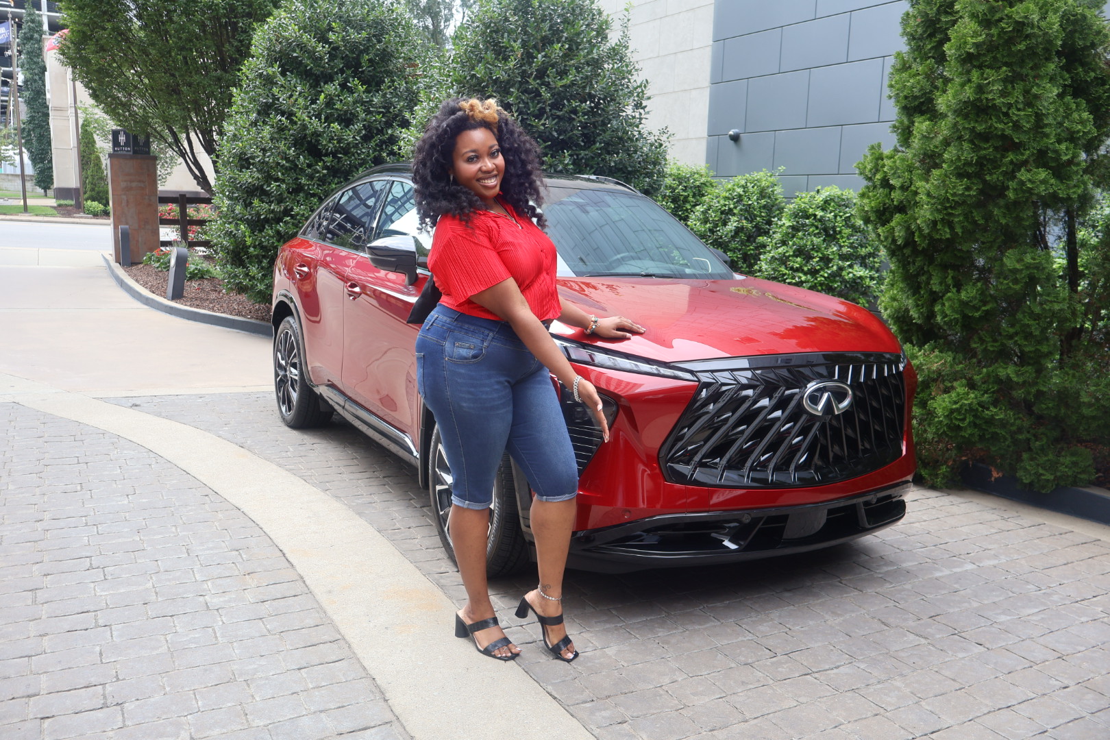 A smiling woman in a red top and jeans stands next to a red Lexus vehicle with a distinctive grille design.