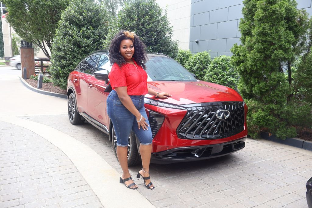 A smiling woman in a red top and jeans stands next to a red Lexus SUV in an outdoor setting with trees and buildings.