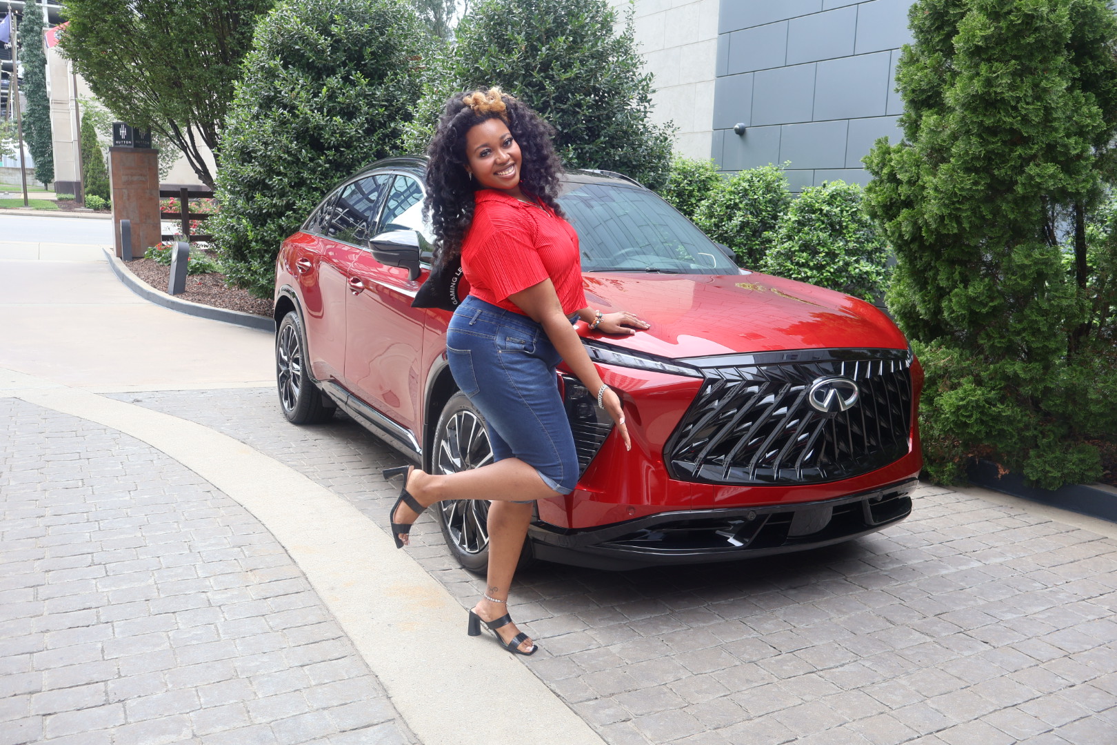 A smiling woman in a red top and jeans stands next to a red Infiniti QX60 SUV in a landscaped outdoor setting.