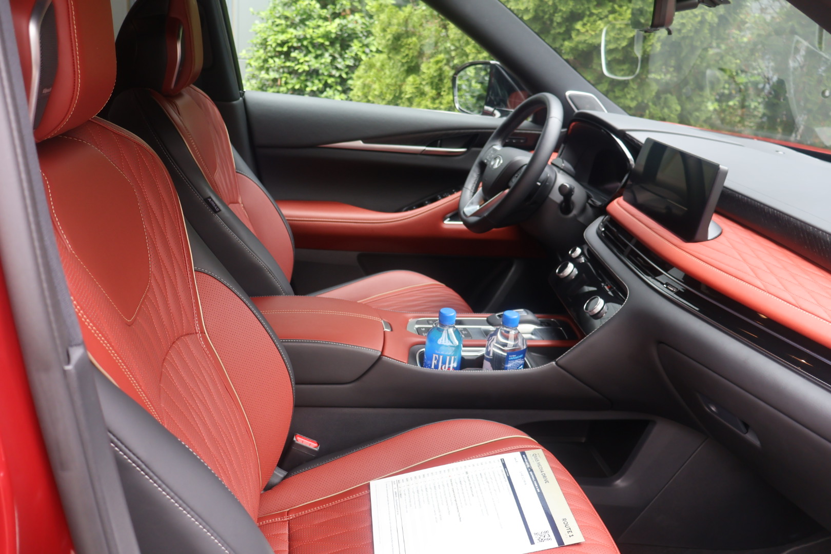 The interior of a red luxury vehicle, with a dashboard, steering wheel, and red leather seats visible. A bottle of water and some papers are on the center console.