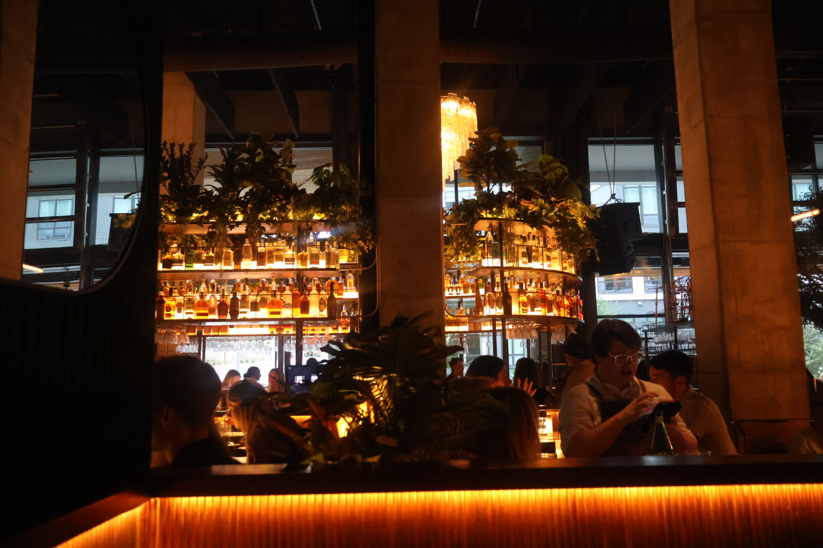 A dimly lit bar interior with shelves of liquor bottles illuminated by warm lighting, surrounded by patrons seated at tables.