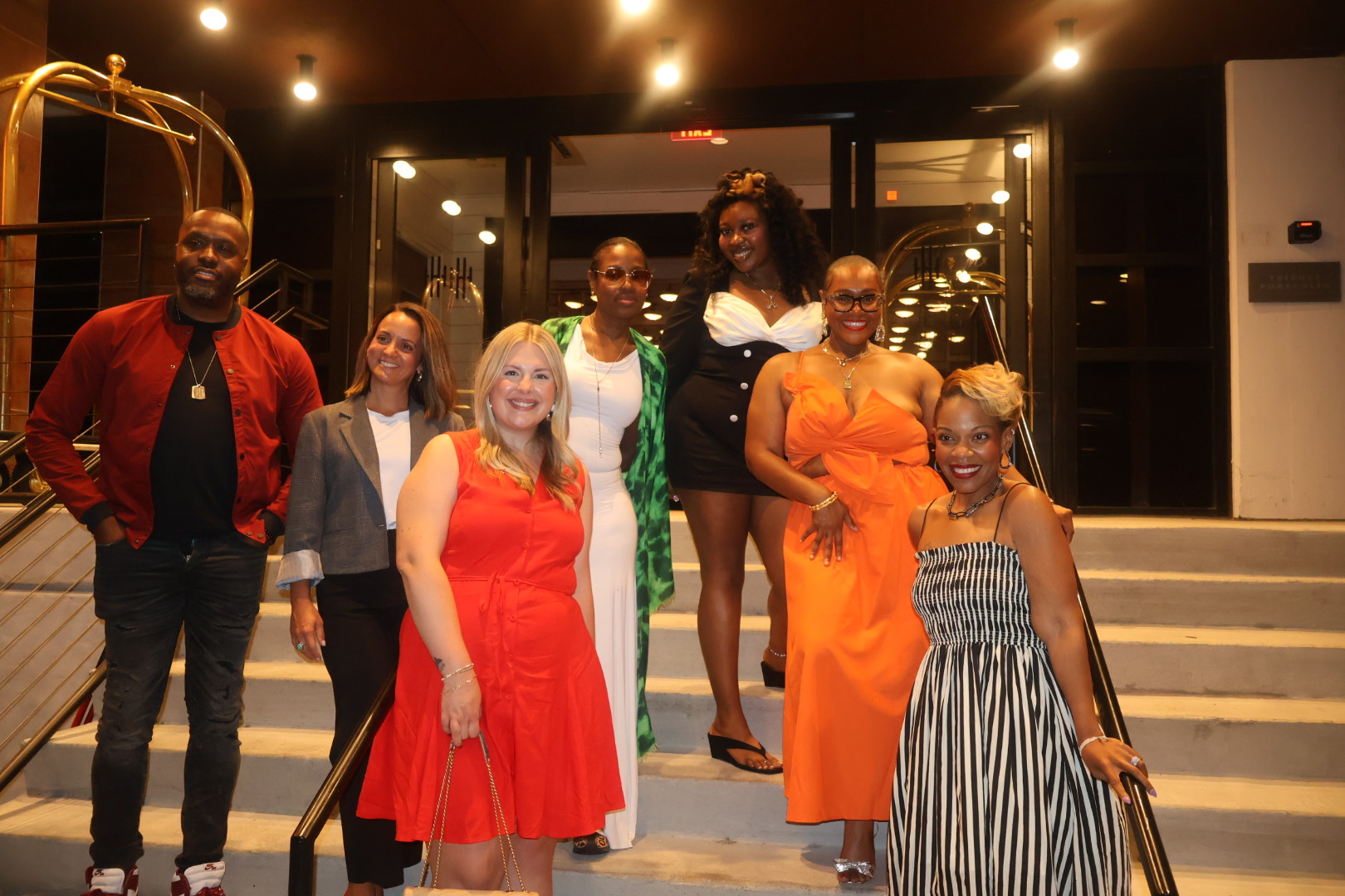 A group of people, some wearing colorful outfits, standing on steps in front of a building with illuminated signage.