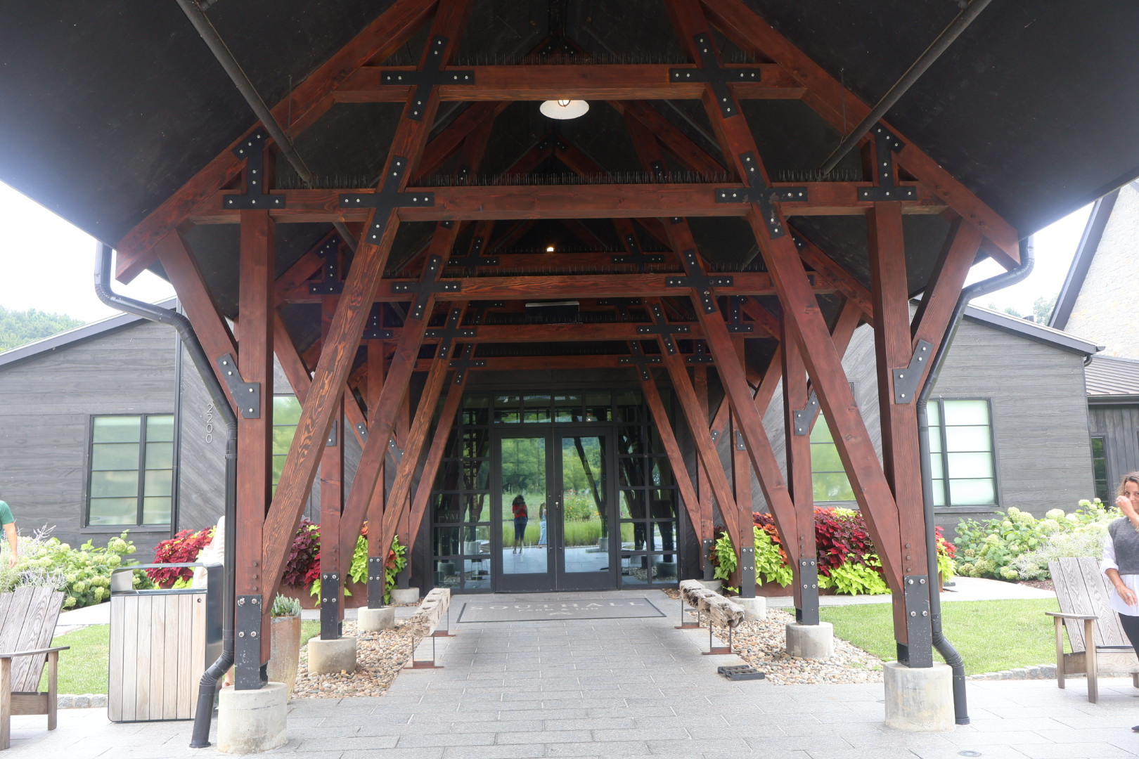 A wooden, truss-style structure with a peaked roof serves as the entrance to a building, surrounded by lush greenery and flowers.