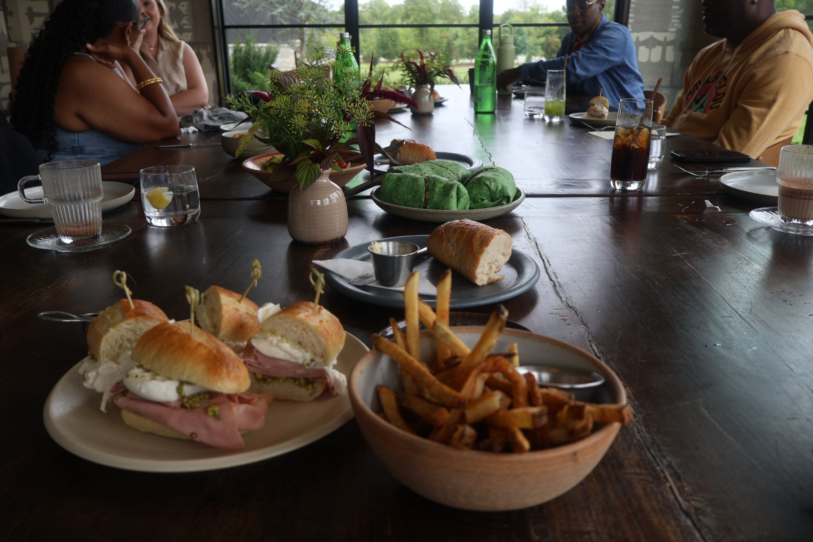 A table set with a variety of food items, including sandwiches, fries, and a green dish, in a restaurant setting with people visible through a window.