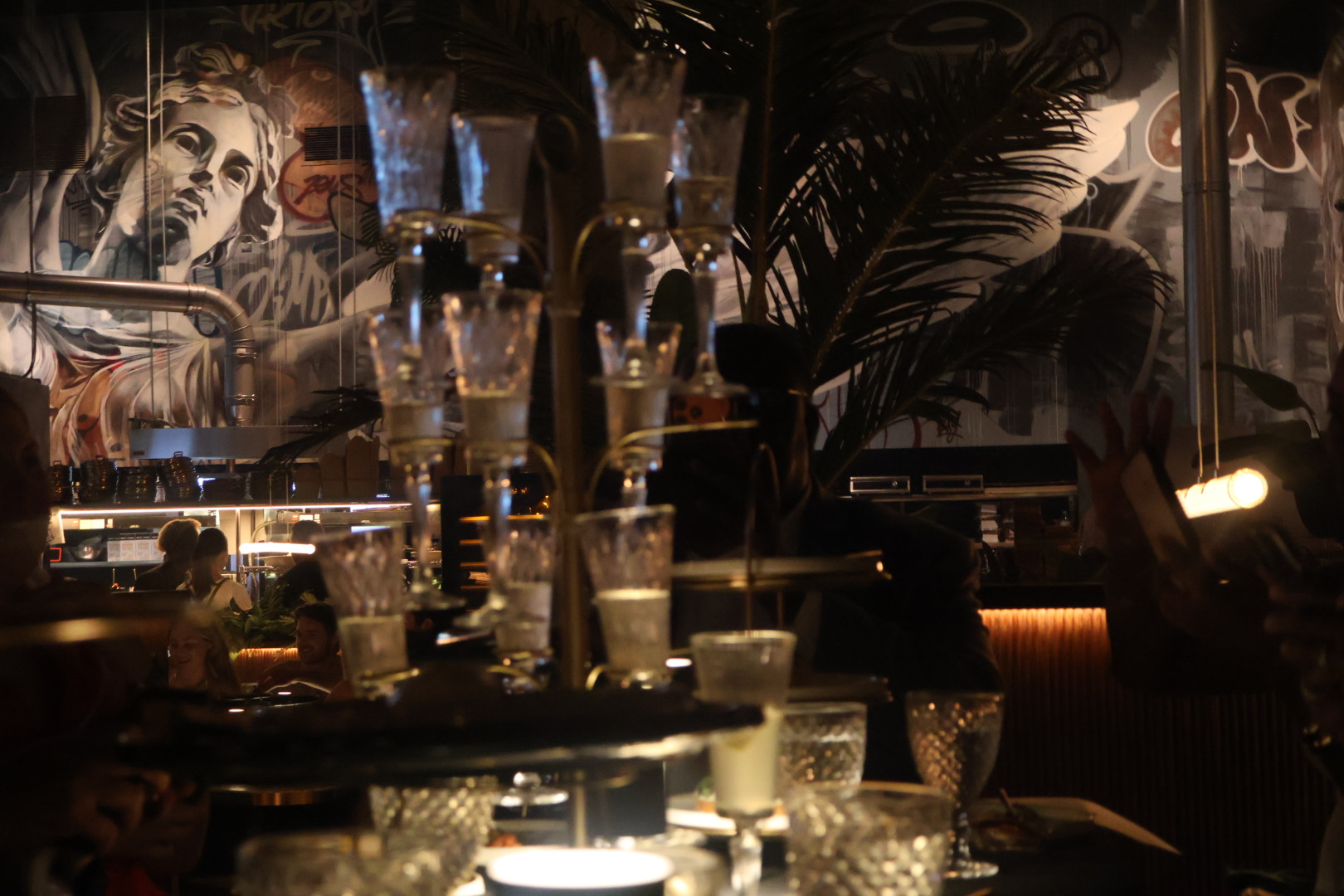 A dimly lit bar or lounge with various glassware, liquor bottles, and a large mural of a woman's face on the wall.