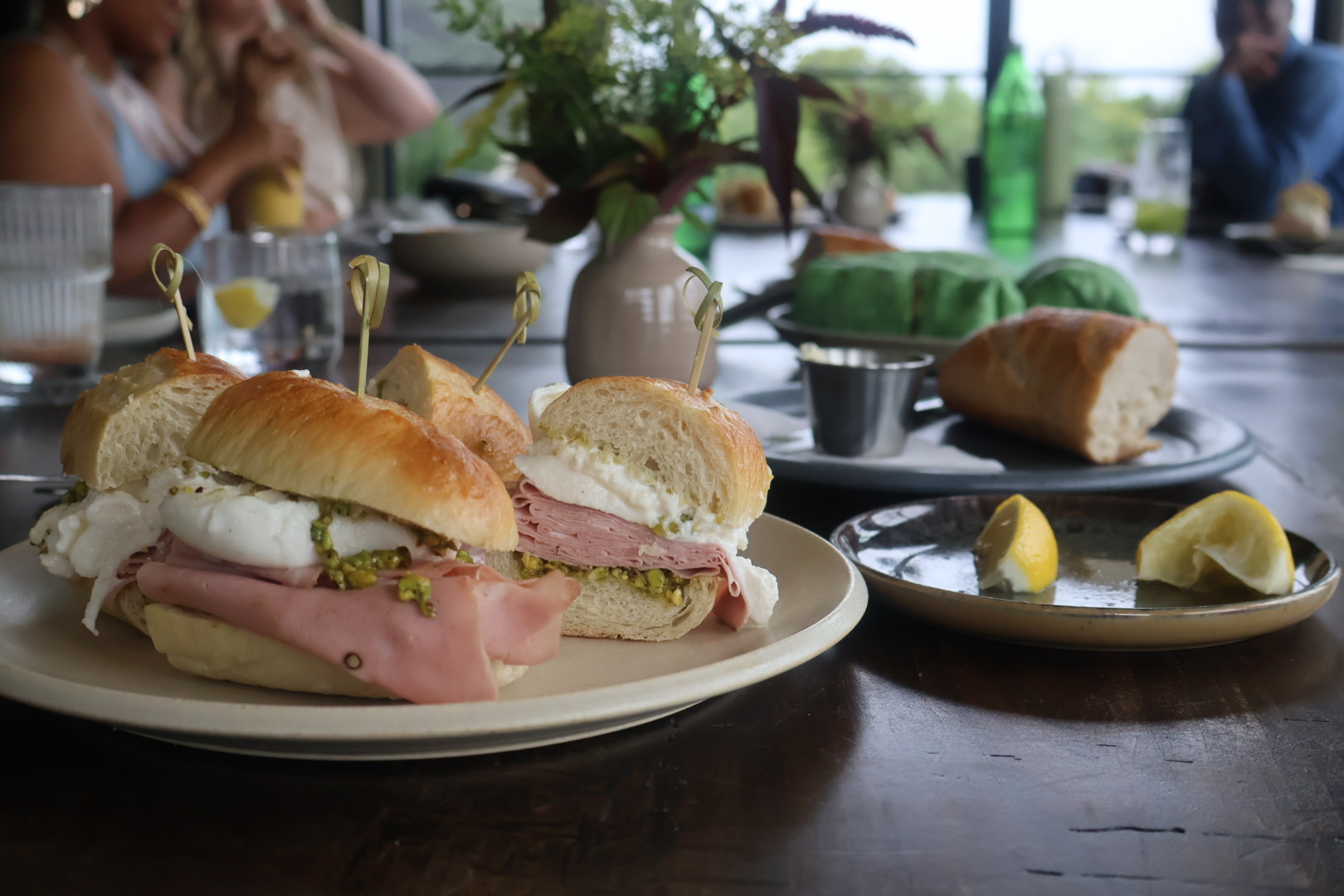 A plate of sandwiches with ham, cheese, and pickles, accompanied by lemon wedges and other side dishes, set on a table with a potted plant and other tableware.
