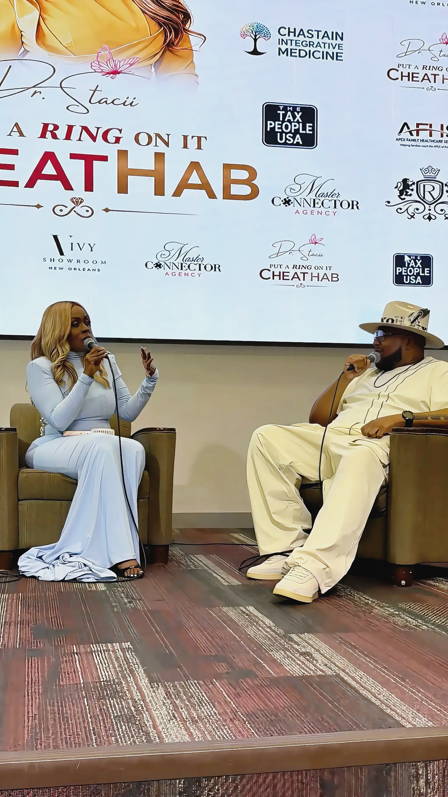Two people sitting on stage, one wearing a white dress and the other wearing a light-colored outfit. Behind them is a large screen displaying text and logos related to "Chastain Integrative Medicine", "Tax People USA", and "Put a Ring on It Cheat Hab".