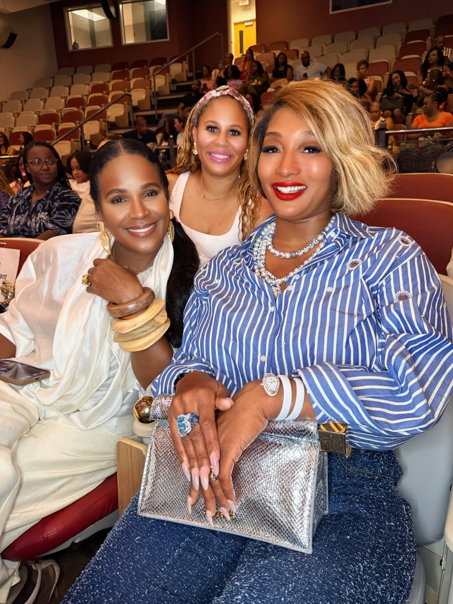 Three smiling women posing together in a theater setting, wearing stylish outfits with bold colors and patterns.