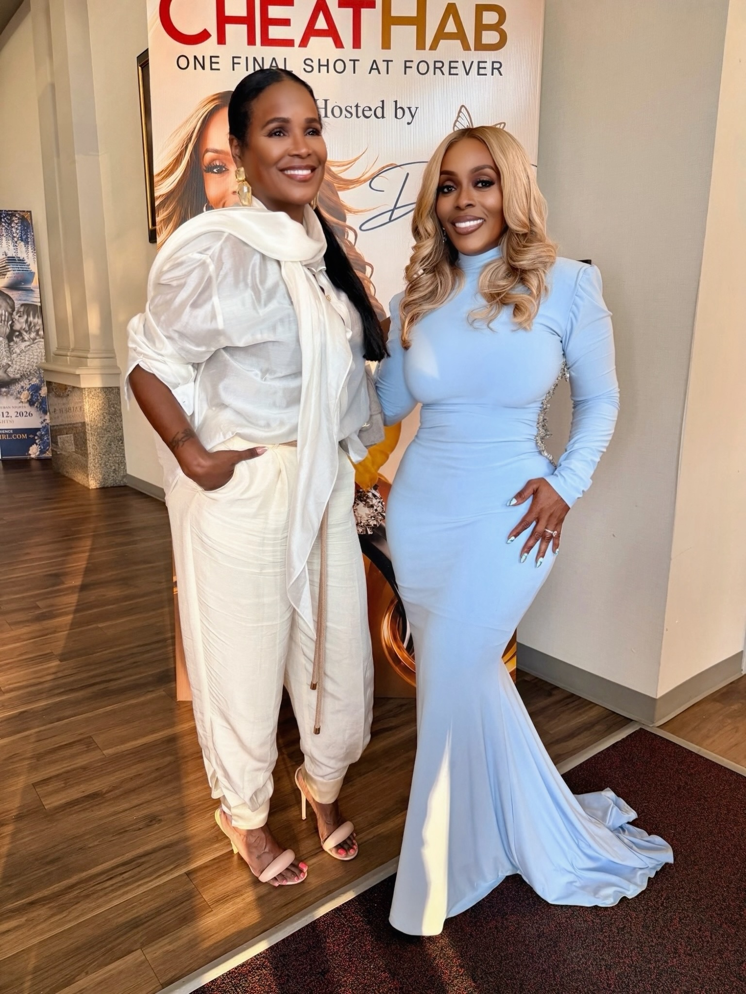 Two women posing in front of a "Cheat Hab" event banner, one wearing a white outfit and the other a blue dress.
