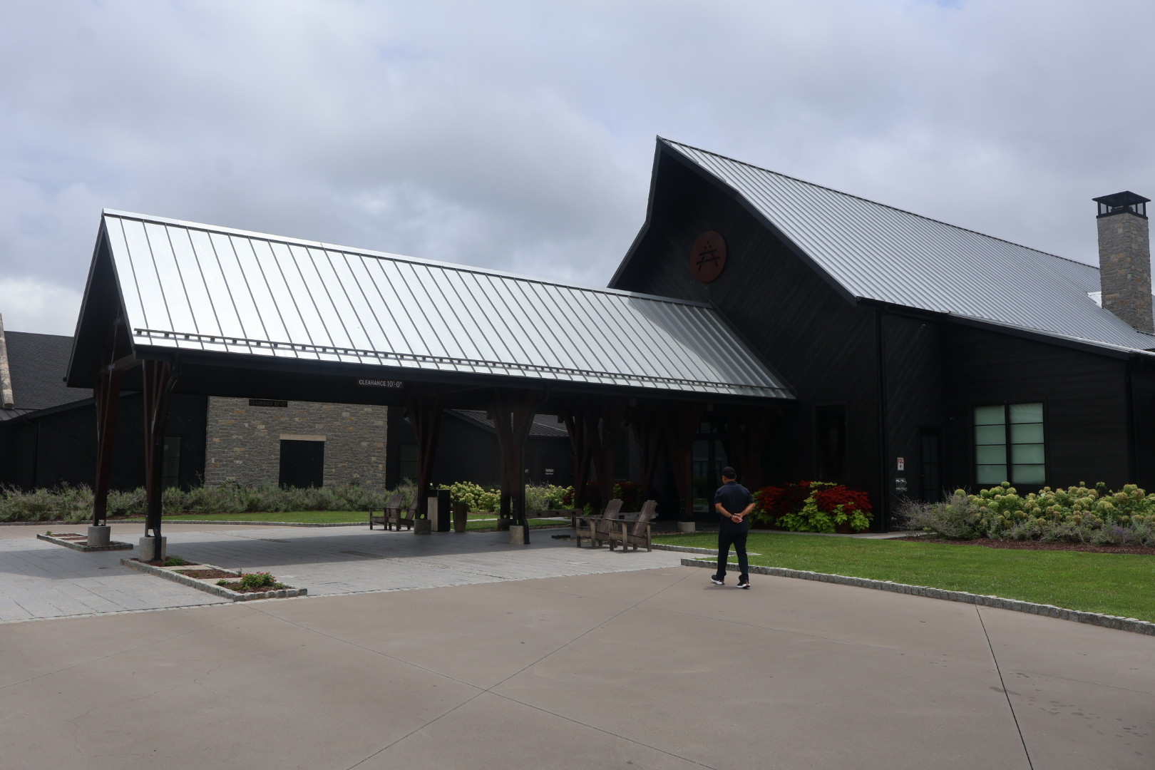 A modern, black-roofed building with a covered patio area and landscaped grounds. A person is standing in the foreground.