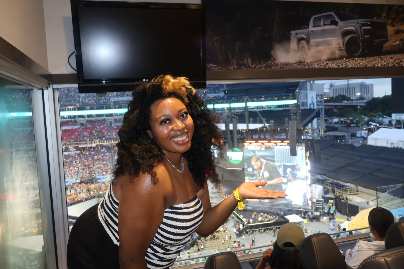 A smiling woman with curly hair stands in front of a large screen showing a sports stadium and a truck advertisement.