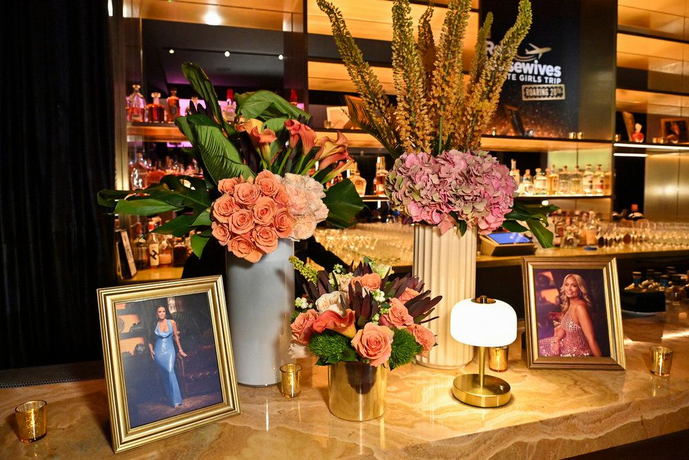 A floral arrangement with roses, hydrangeas, and greenery displayed on a bar counter, along with framed photos and a lamp. The Bravo logo is visible on the wall behind the display.