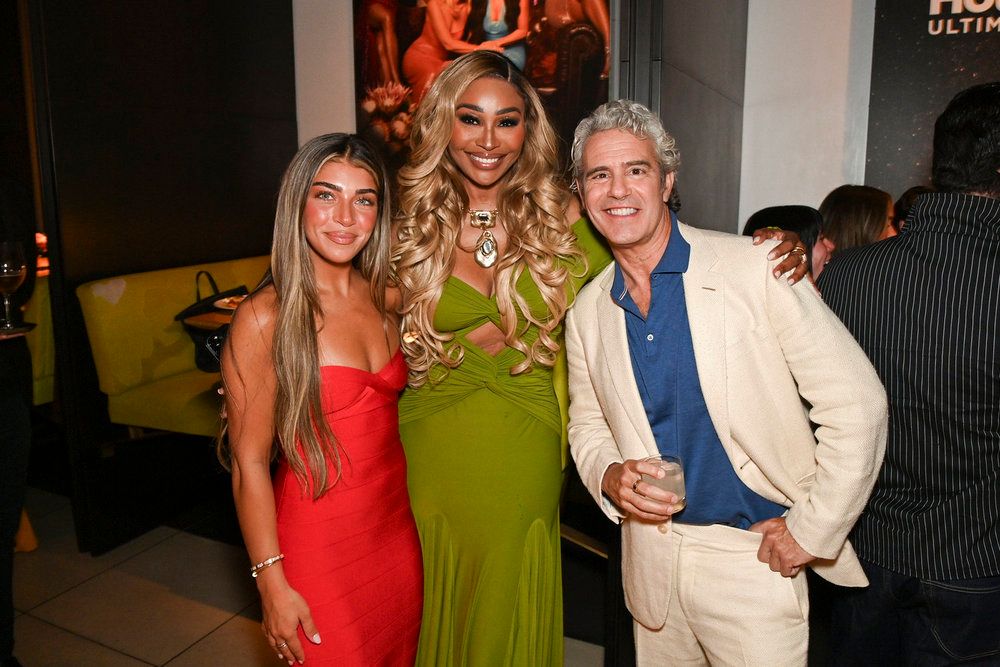 Three women in formal attire posing together, including a woman in a green dress and two other women in red and white dresses, with a man in a light-colored suit standing with them.
