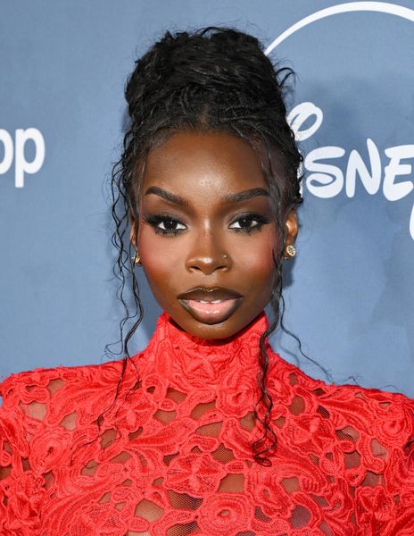 A woman with dark skin and braided hair wearing a red lace dress posing at a Disney+ event.