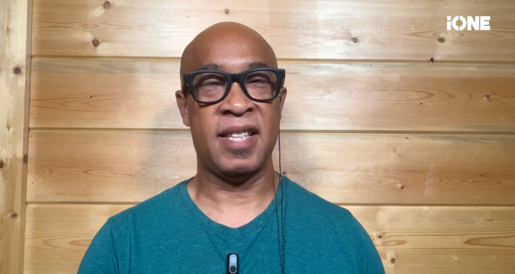 A bald man with glasses wearing a teal shirt, standing in front of a wooden wall.