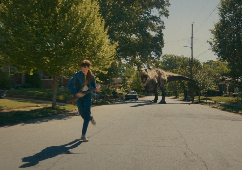 A person running down a sidewalk in a park, with large dinosaur statues in the background.