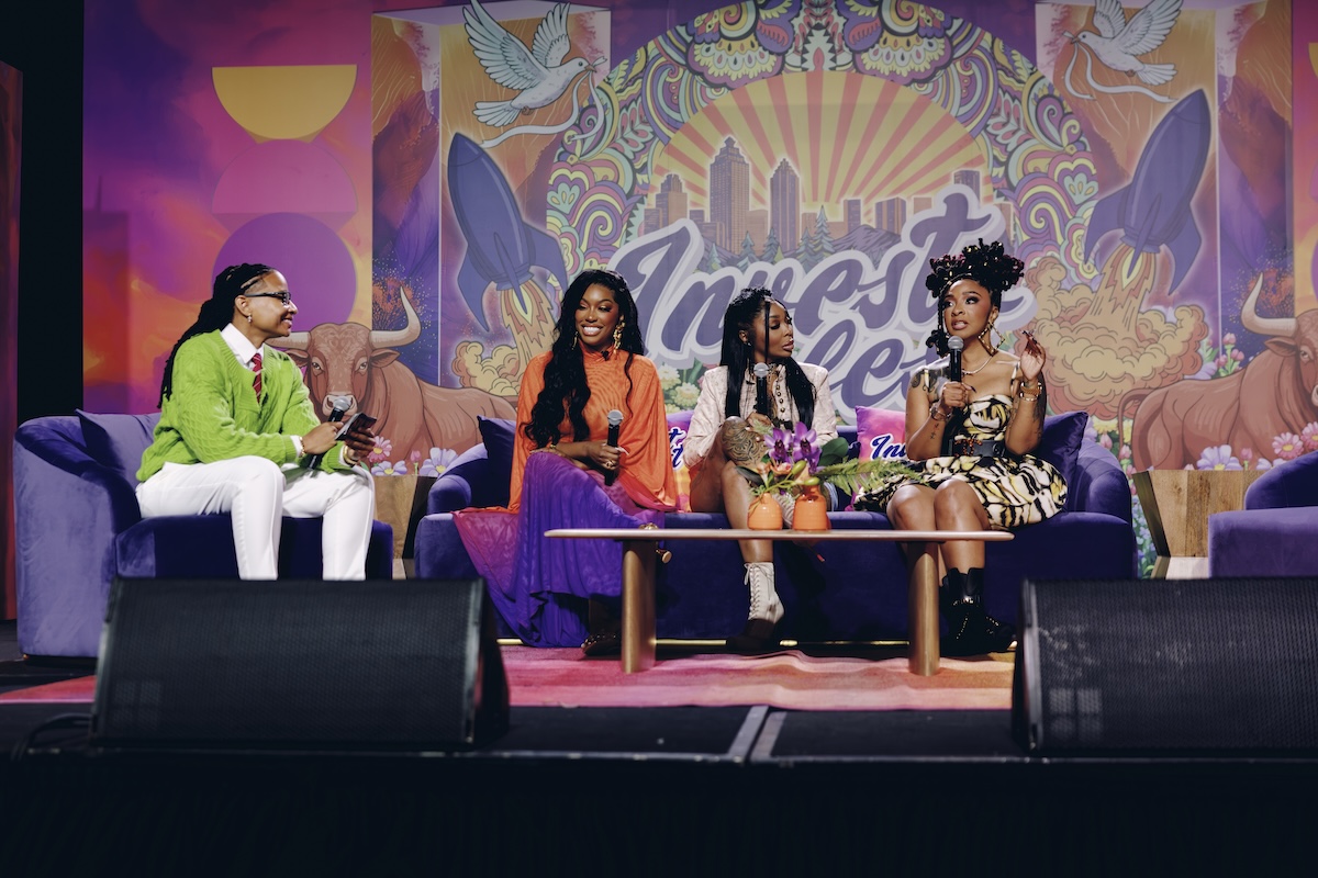 Four people sitting on a colorful, patterned stage set with a cityscape backdrop and winged figures.