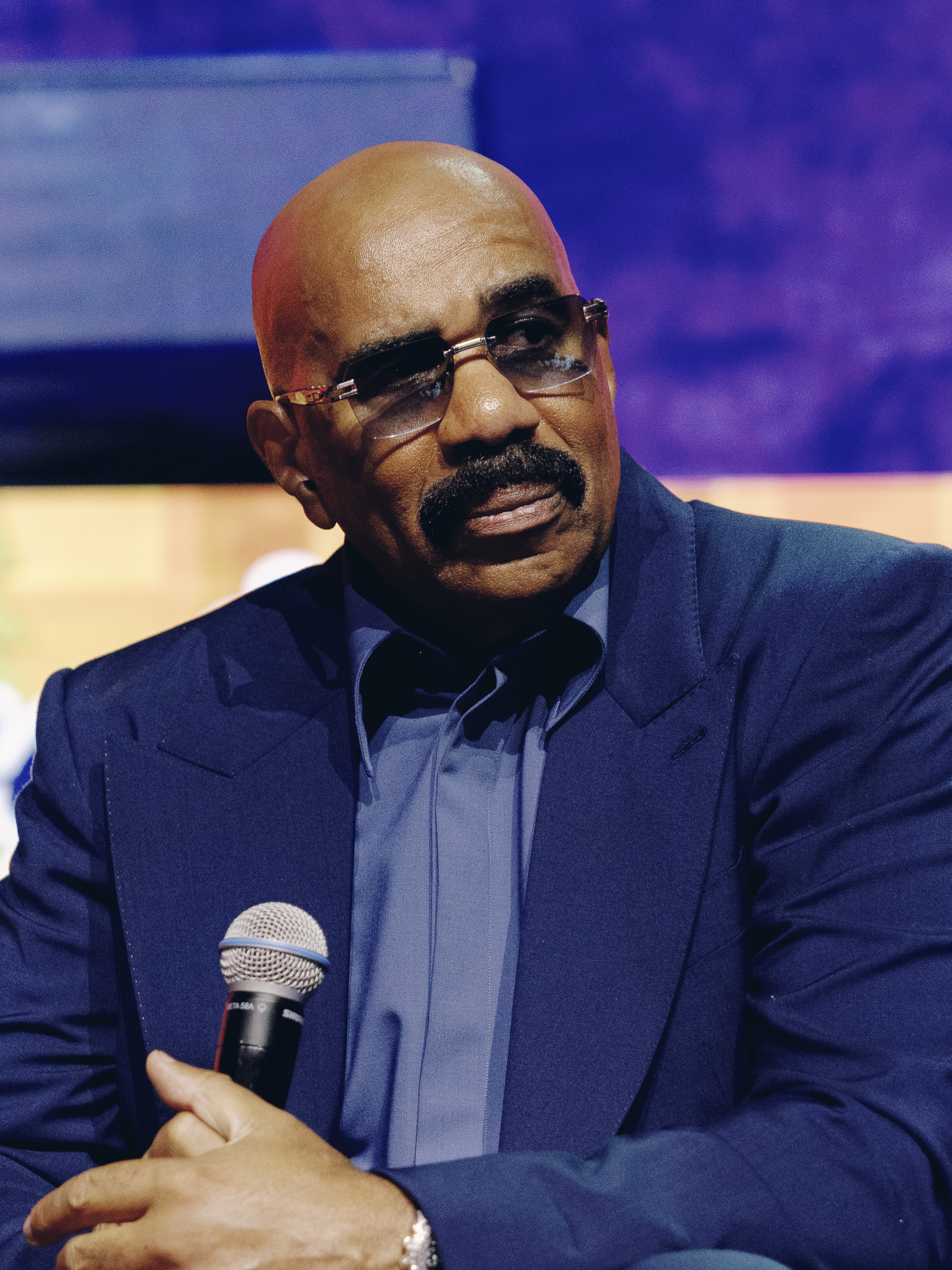 A bald, middle-aged Black man wearing glasses and a dark suit, holding a microphone and speaking at a podium against a purple and blue background.