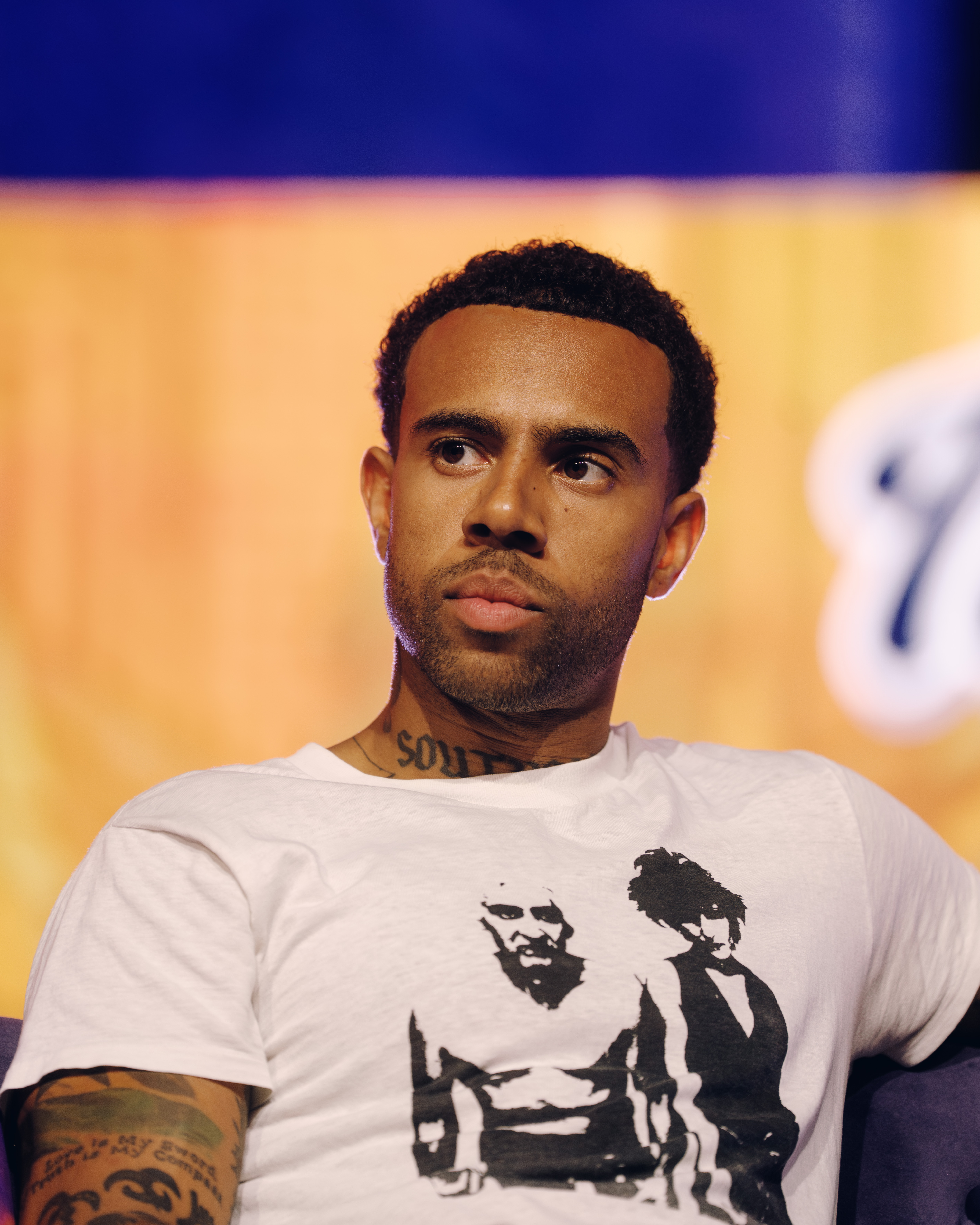 A man with a beard and tattoos wearing a white t-shirt with a graphic design, standing against a blue and yellow background.