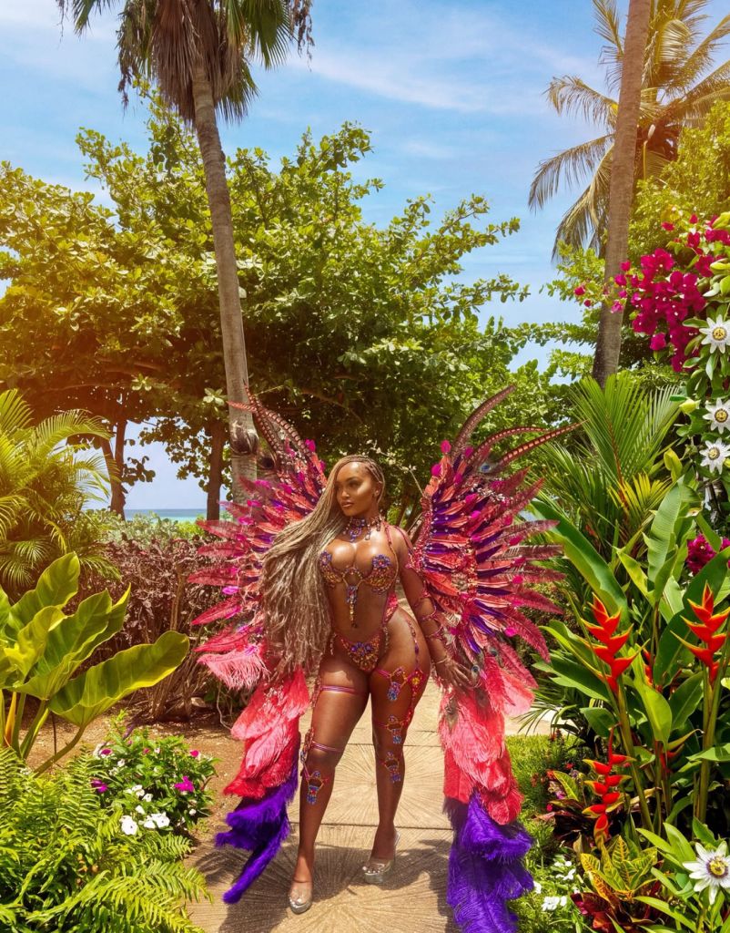 A woman wearing a colorful, feathered costume stands in a lush tropical garden with palm trees, flowers, and greenery.