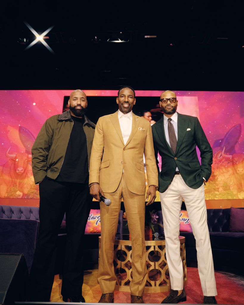 Three men in formal attire standing on a stage with colorful background and lighting.