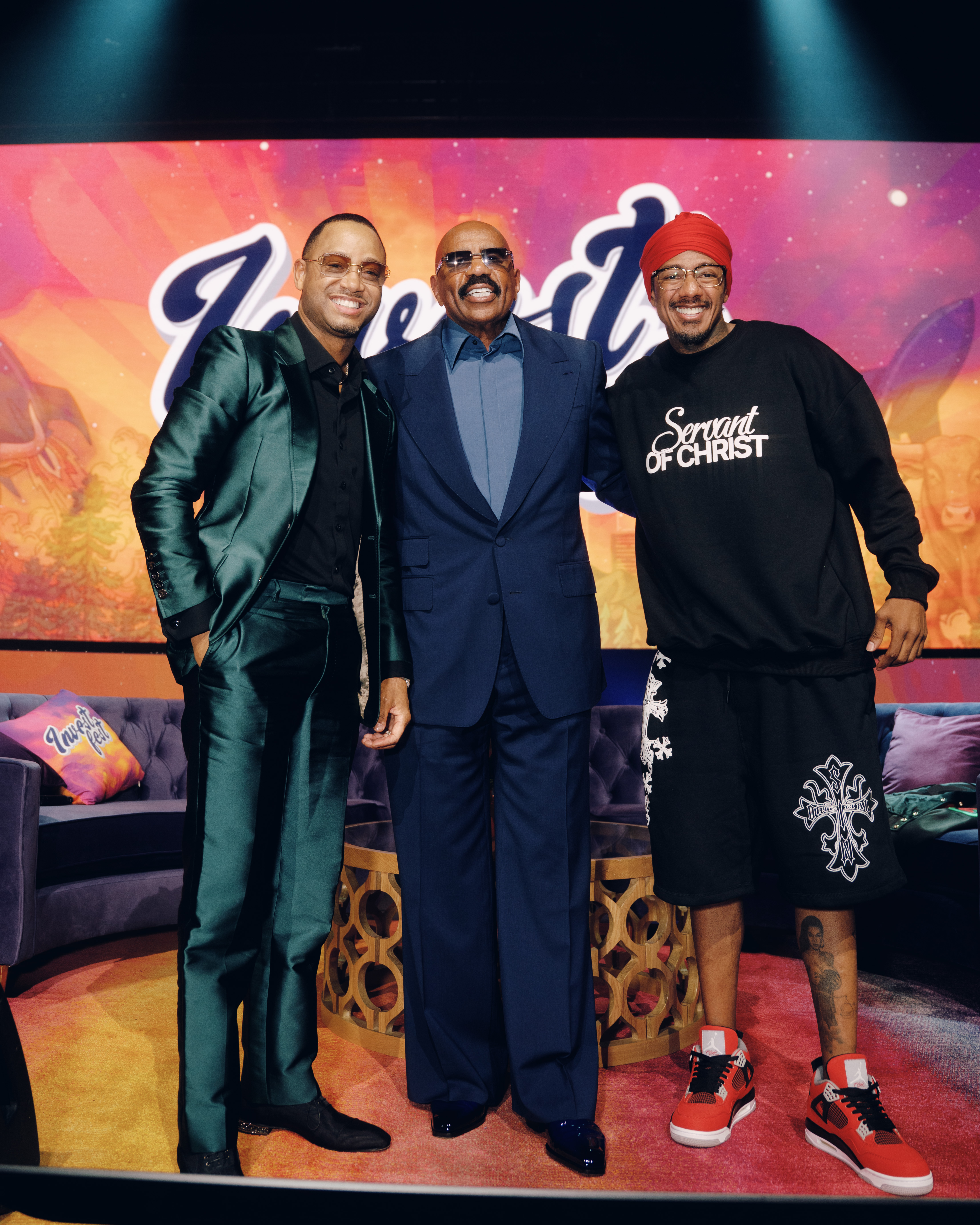 Three men posing together on a colorful stage, one wearing a "Servant of Christ" sweatshirt.