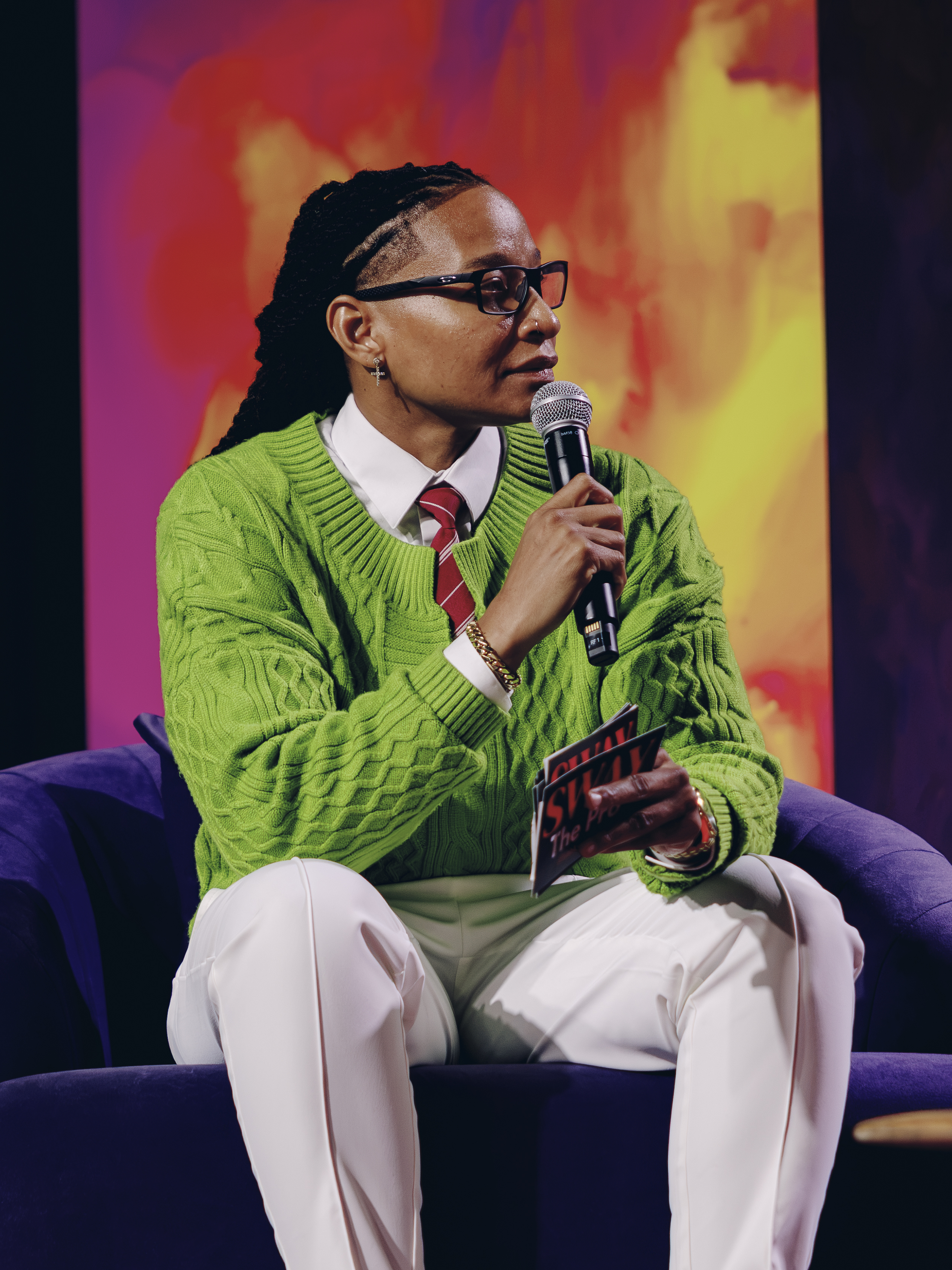 A person wearing a green sweater and glasses speaking into a microphone on a purple couch against a colorful abstract background.
