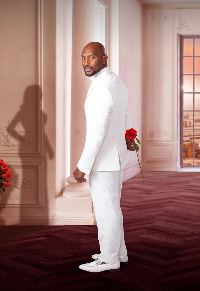 A well-dressed man in a white suit stands in a formal room, holding a red rose.