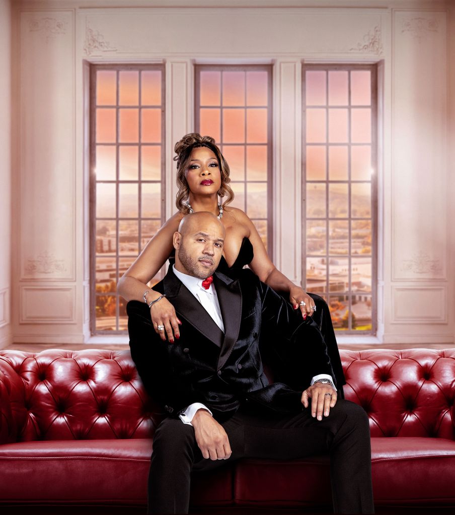 A well-dressed couple posing in a luxurious room with large windows overlooking a cityscape at sunset.
