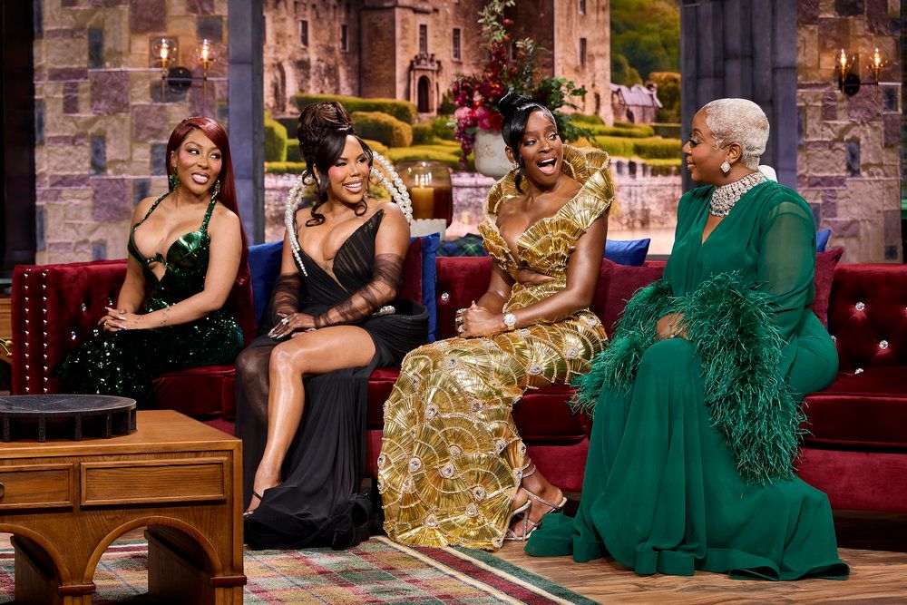 Four women in glamorous dresses sitting on a couch in front of a castle-like building.