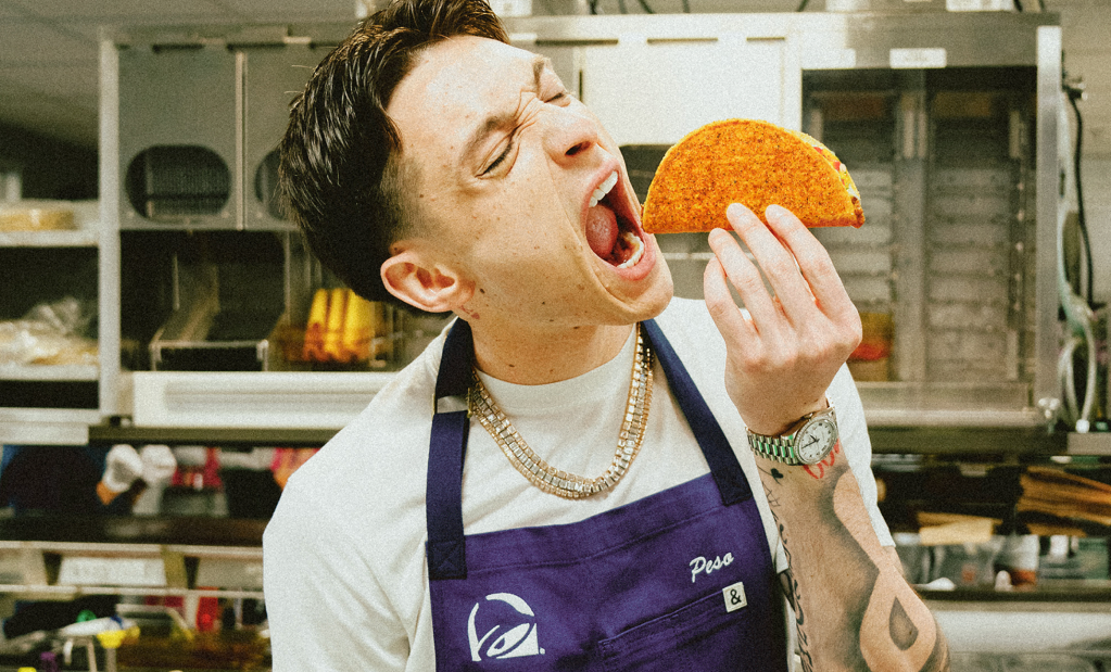 A person wearing a purple apron and jewelry enthusiastically bites into an orange taco shell.