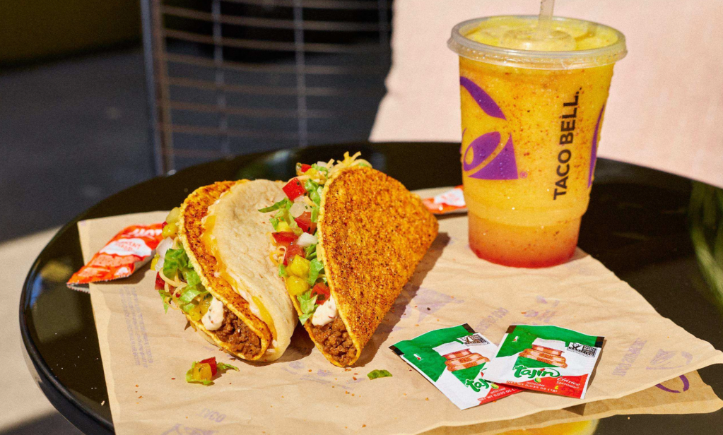 Two tacos with ground beef, lettuce, and tomatoes on a black tray, accompanied by a Taco Bell branded drink cup.