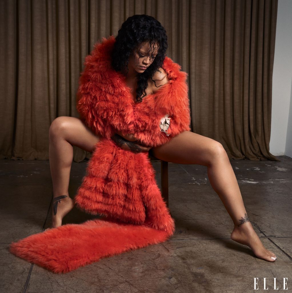 A person wearing a bright red furry coat sitting on the floor with their legs crossed, looking down.