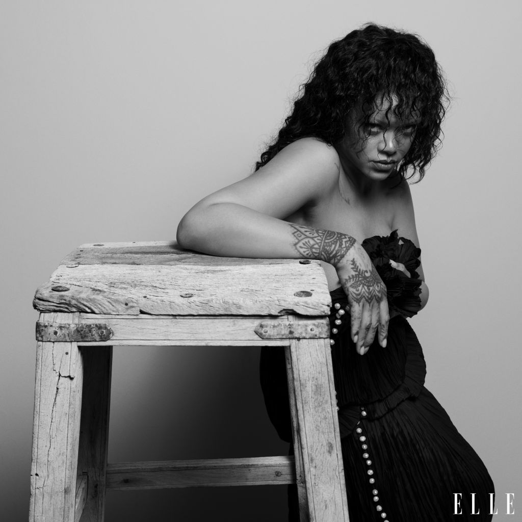 A Black and white portrait of a woman with curly hair and tattoos, sitting on a wooden bench with a pensive expression.